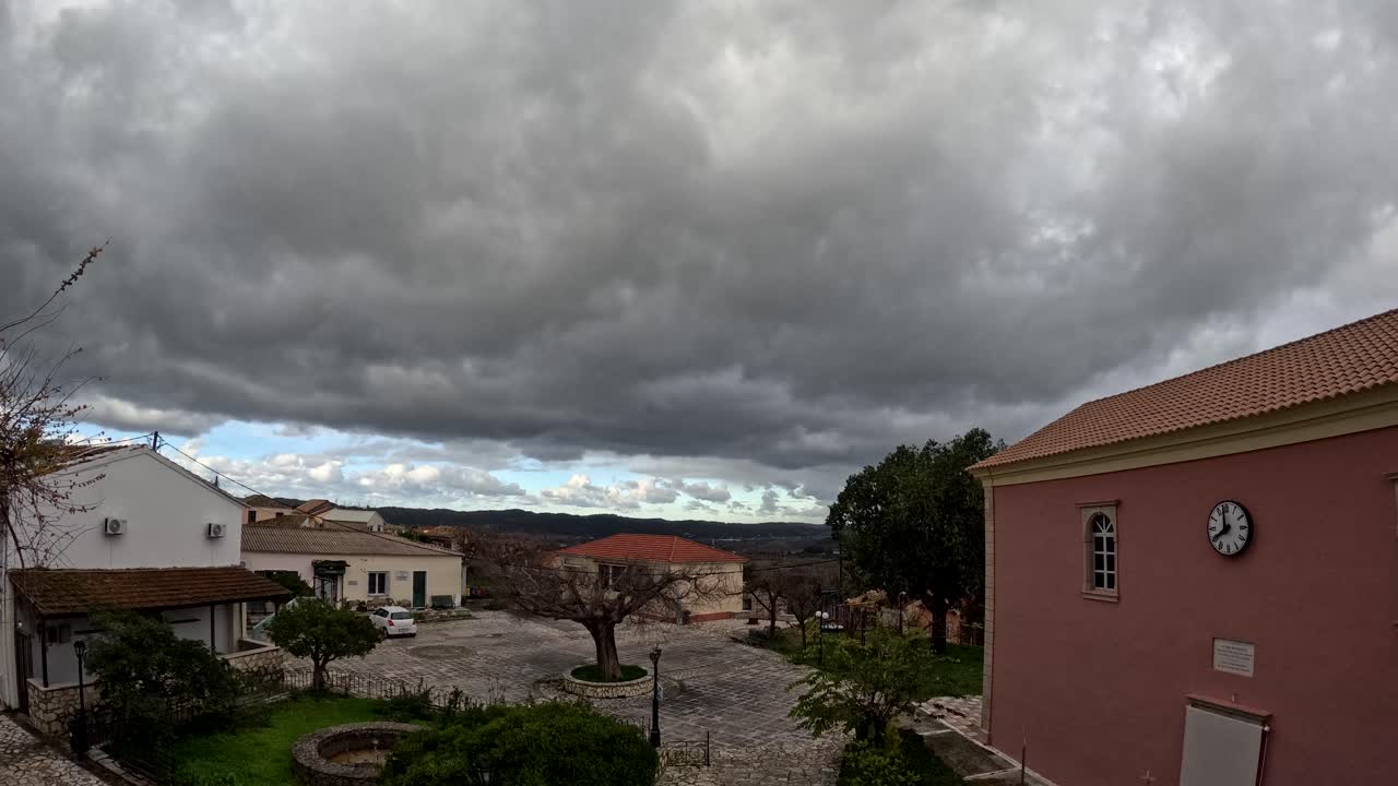 Timelapse panoramico di nuvole spettacolari che passano sopra una piazza tradizionale di un villaggio greco con una chiesa rosa con torre dell'orologio e pavimentazione in pietra a Corfù, in Grecia, che mette in mostra l'architettura e la vita rurale