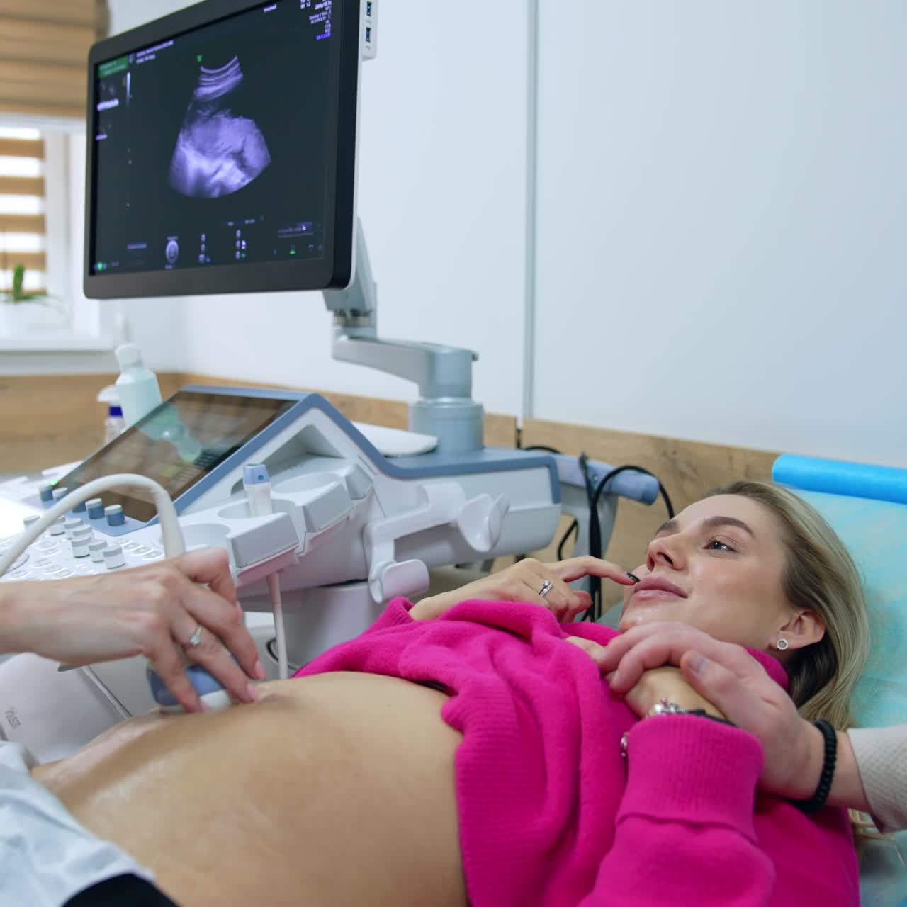 Pregnant woman in pink sweater is examined by the doctor on ultrasonic equipment. Caucasian couple expecting a child at ultrasound check up