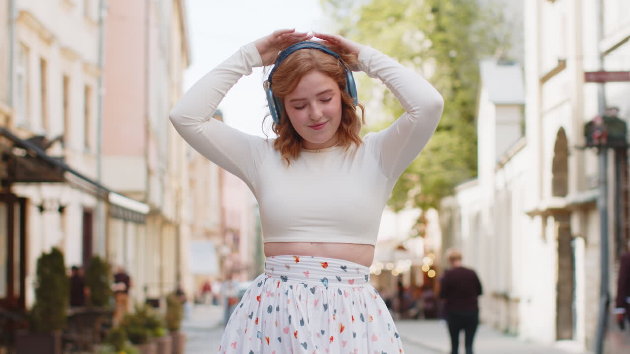 feliz mujer pelirroja sonriente en auriculares inalámbricos escuchando música bailando al aire libre en la calle de la ciudad