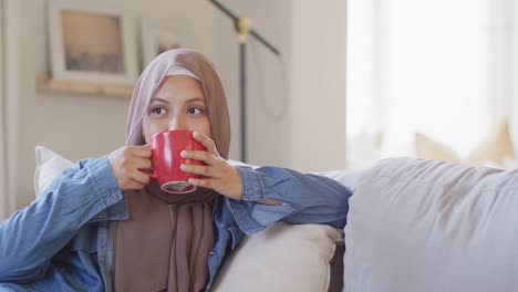 Video-of-smiling-biracial-woman-in-hijab-drinking-coffee-sitting-in-living-room-at-home