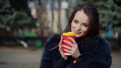 fashion outdoor video of beautiful sensual girl with dark hair wearing luxurious fur coat,holding paper cup with coffee.