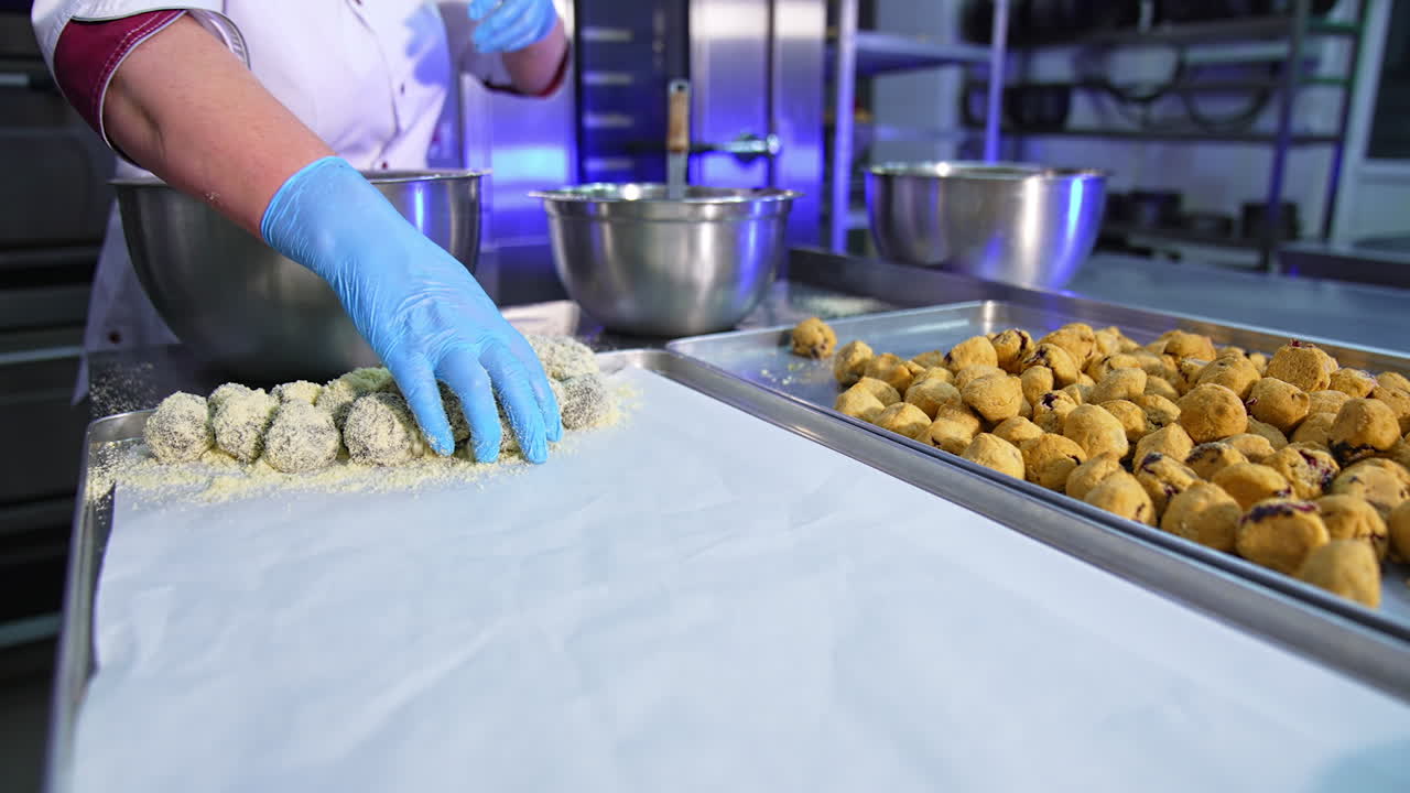 Confectionery worker covers the biscuits with sprinkling. Female hands take the sweets, puts them into a metal bowl, mixes and puts them on a tray.