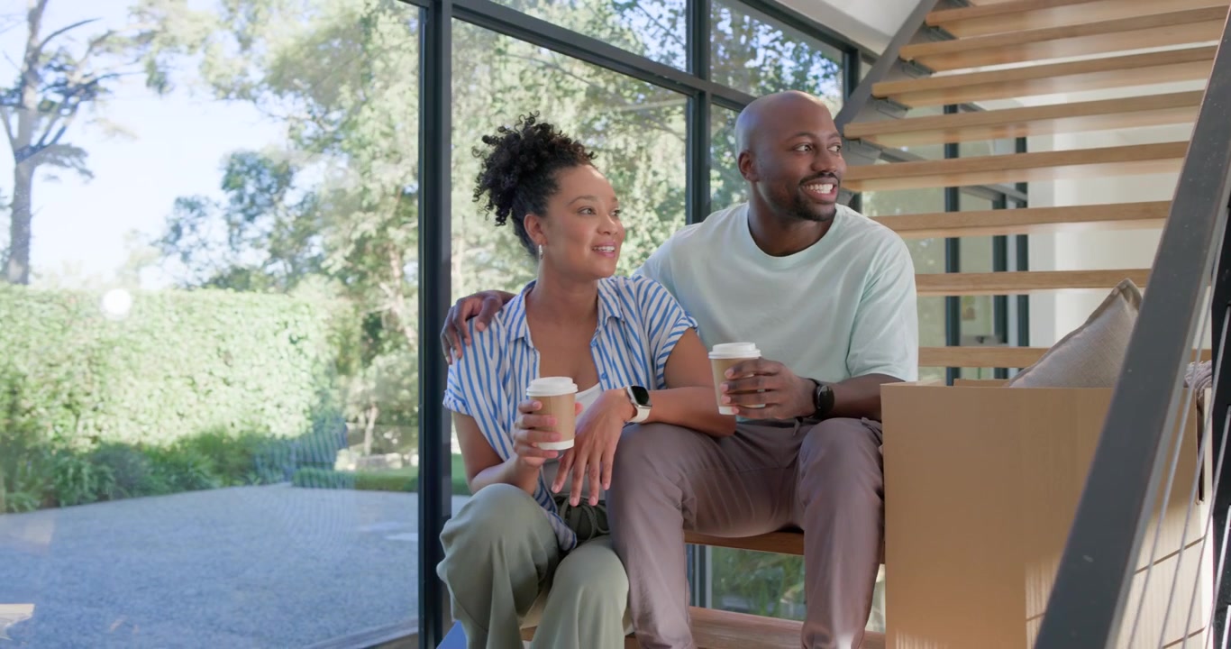 African American couple on stairs by glass wall holding cups, man placing arm, pointing amid boxes