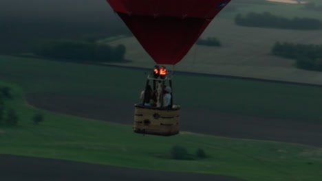 three friends in moving hot air balloon drifting over green farmland at dusk, burner flames glowing above wicker basket, aerial view capturing serene travel adventure