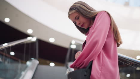 joven vestida con un vestido rosado y una camiseta negra en una escalera mecánica, buscando en su bolsa negra, pareciendo reflexiva, la escalera se mueve hacia arriba mientras ella revisa su bolsa con concentración