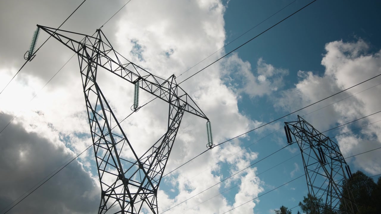 Steel lattice power pylons and suspended lines rise against a bright, fast-moving cloudy sky. Strong visual for energy, infrastructure, or environmental themes.