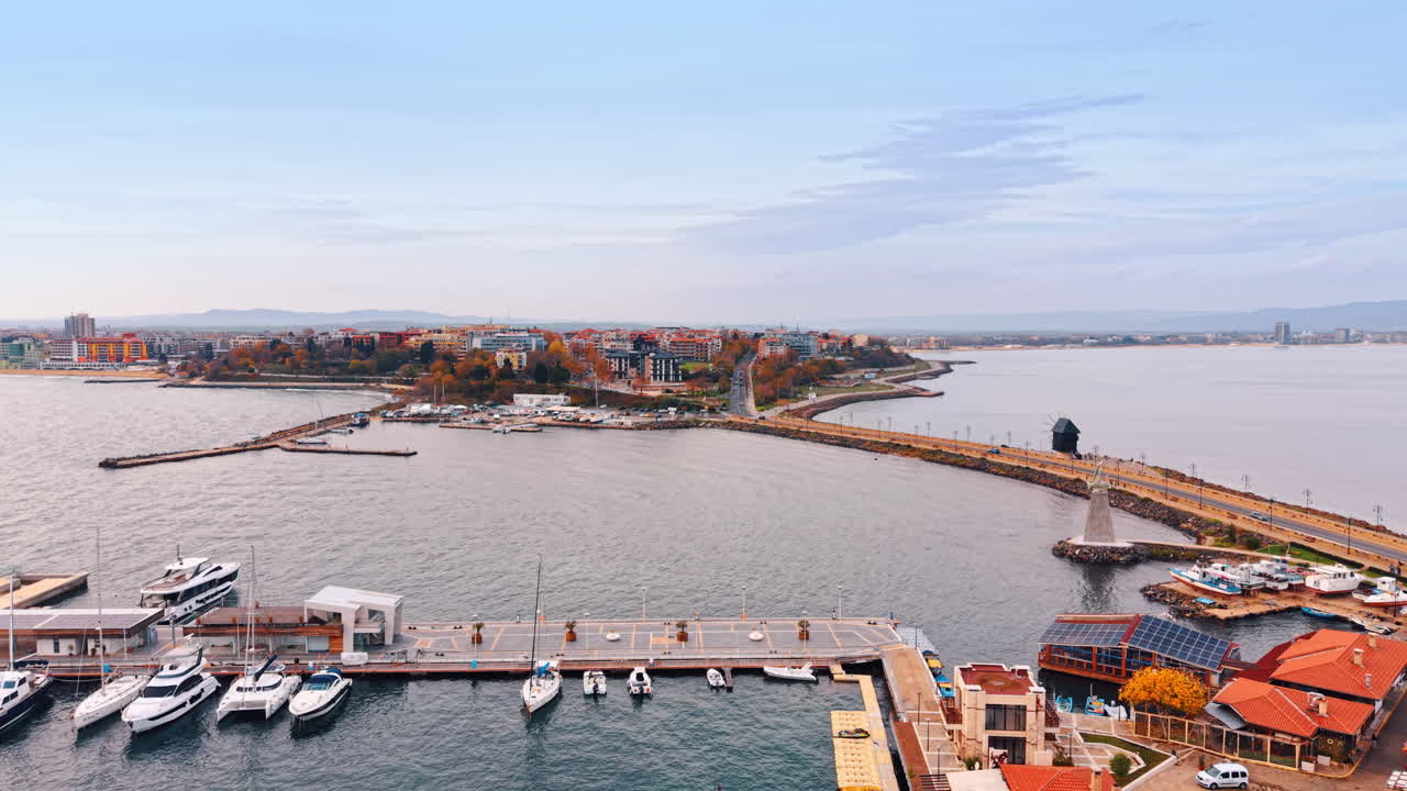 Volo sopra le case dai tetti rossi, la banchina con le barche e lo specchio d'acqua grigio. Avvicinamento alla diga con la strada che conduce alla penisola. Nessebar, Bulgaria.