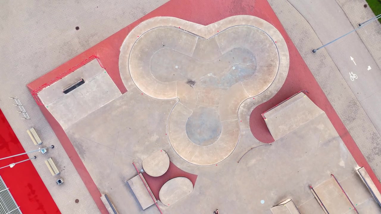 Top-down drone view of a concrete skatepark with flowing bowls, ramps and red perimeter paving beside benches and a marked cycle path.