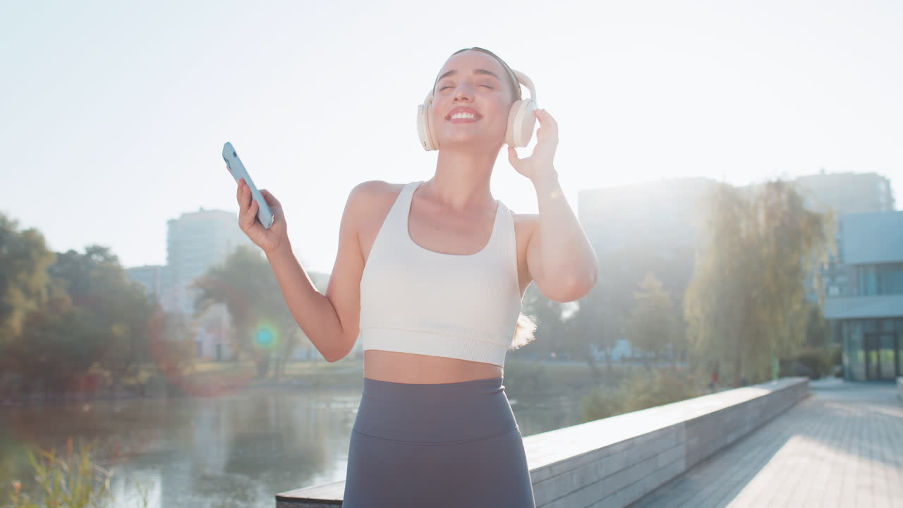 행복한 젊은 여성 조거가 헤드폰과 스마트폰을 통해 음악을 듣고 도시 공원에서 고 있습니다.