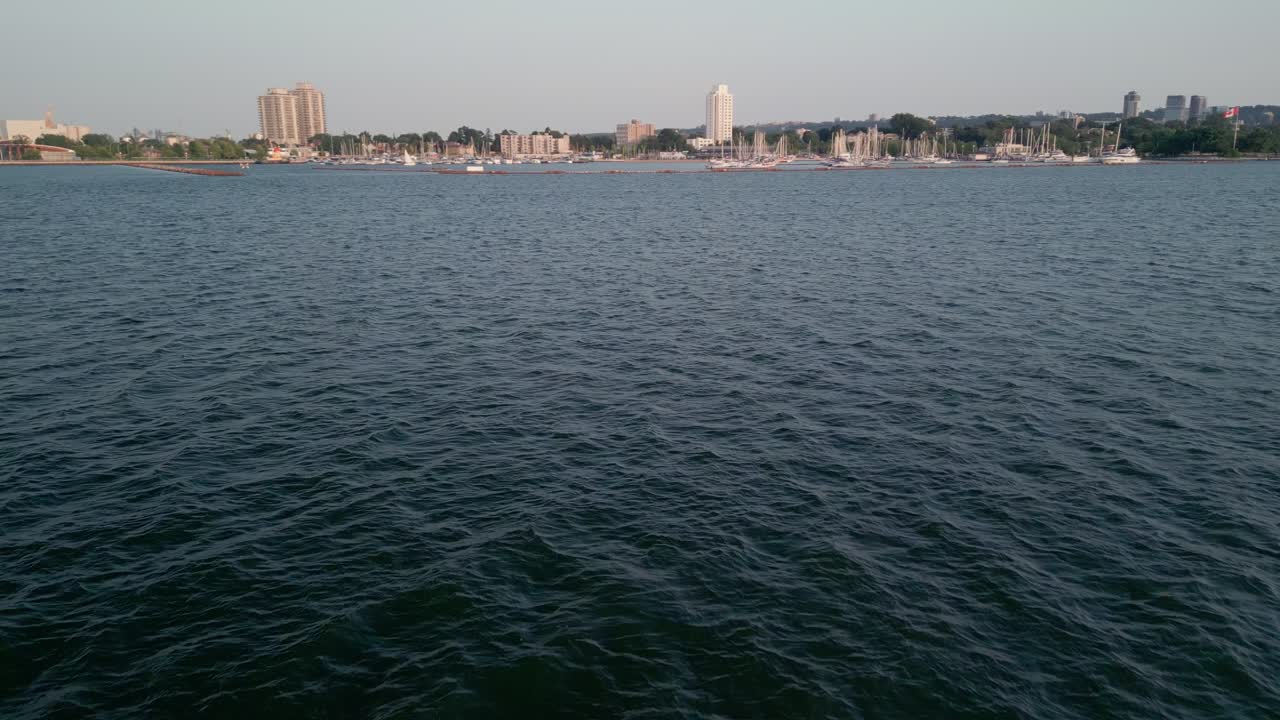 Un drone sorvola le acque blu profonde del Lago Ontario nel porto di Hamilton, catturando alti edifici sullo sfondo.