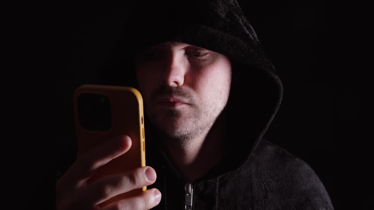 Uomo con una felpa scura con cappuccio che guarda il suo smartphone, rappresentando la dipendenza digitale, l'uso dei social media e il comportamento online.
