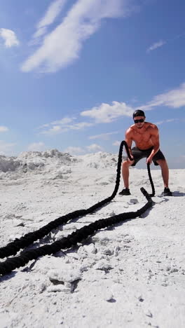 male athlete exercising outdoors around white hills at daytime. active physical workout. strong man in sunglasses has a workout with a rope on the ground. vertical video