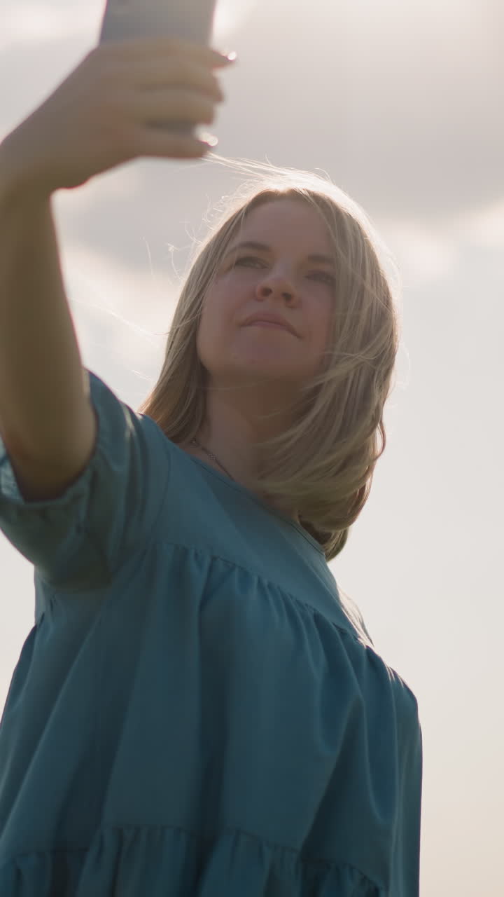 primer plano de una mujer con un vestido azul, sosteniendo un teléfono inteligente mientras se filma al aire libre. toca suavemente su cabello mientras sopla en la brisa, capturando un momento en el campo iluminado por el sol