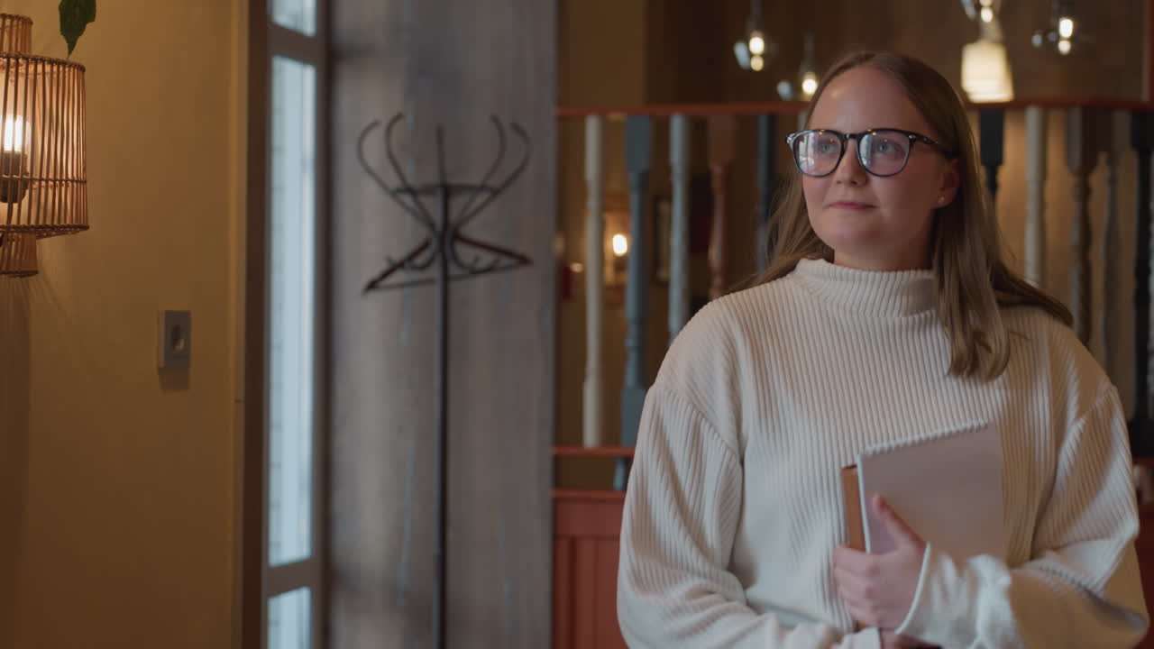 indoor restaurant entrance with warm lighting and vintage decor as excited young lady holding book enters through glass door, cozy ambiance featuring wall lamp, coat rack, and red wooden divider