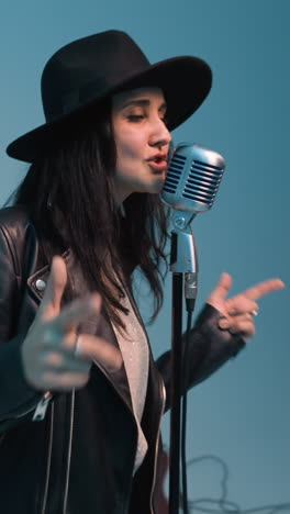 un vocalista con una chaqueta de cuero negra y sombrero, sonriendo y cantando con los ojos abiertos, de pie en un micrófono vintage con las manos moviéndose arriba y abajo un tecladista trabaja un sampler rojo contra un fondo azul