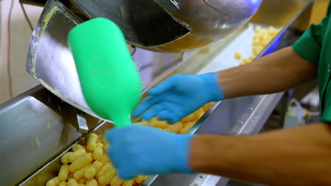 Factory worker takes corn sticks out of a metal tank. Crispy puff corns are put on the conveyor belt for the next stage of production.
