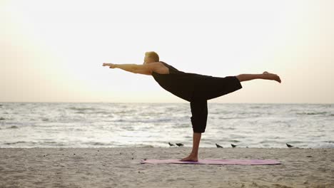 a young man is doing yoga on a sports mat, standing on one leg during the dawn of the son. doing a specific exercise. meditation