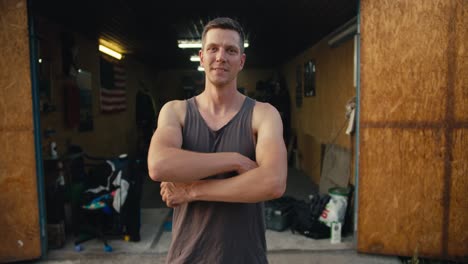 Portrait-of-a-happy-man-with-a-short-haircut-in-a-gray-T-shirt,-who-folds-his-arms-on-his-chest-and-poses-in-front-of-his-garage-workshop.-Happy-mechanic-at-his-favorite-job