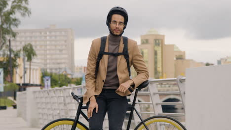 handsome african-american man in formal clothes with helmet and backpack looking and smiling at the camera while leaning on a bicyle on the city bridge