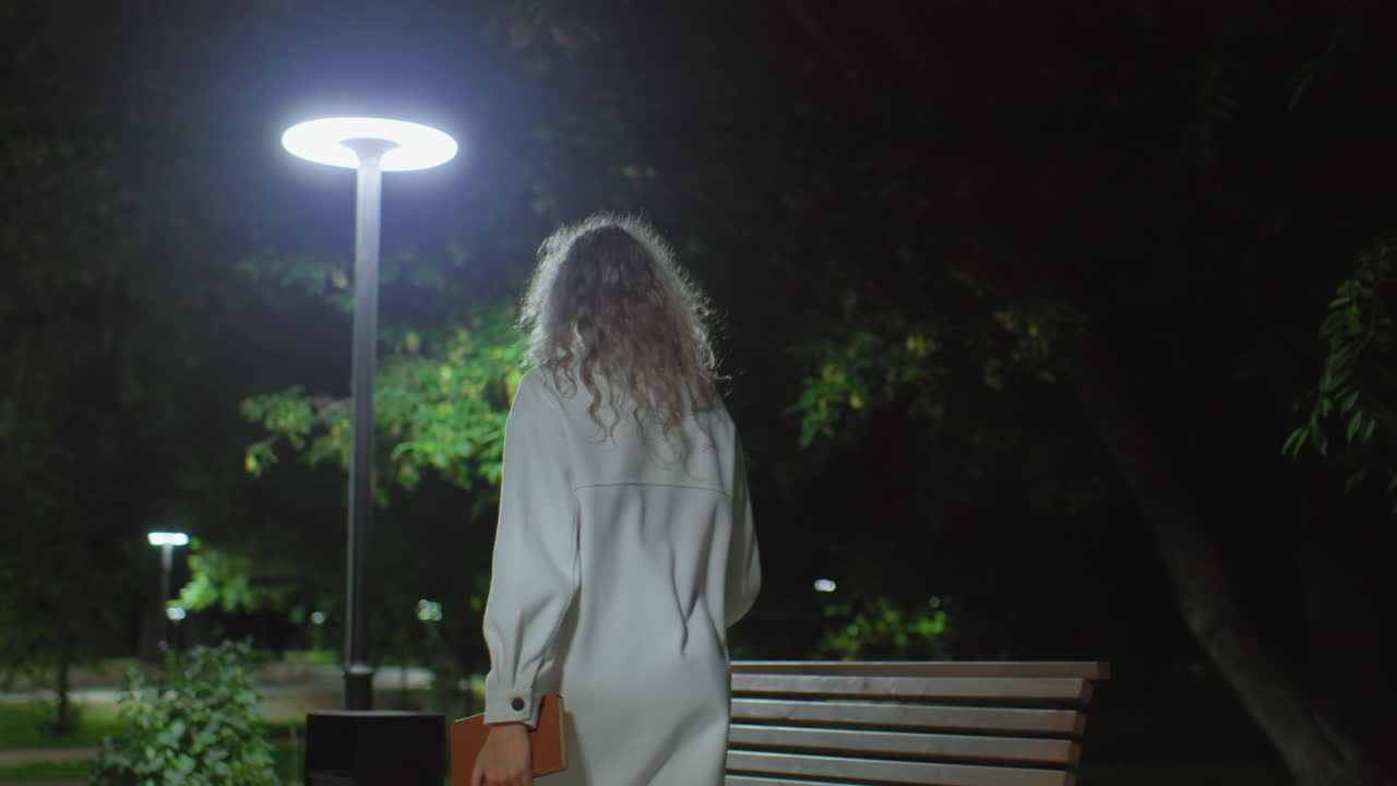 Back view of young lady in white coat standing near wooden bench under bright streetlight, seated outdoor during cool evening while gently opening book to read, surrounded by greenery