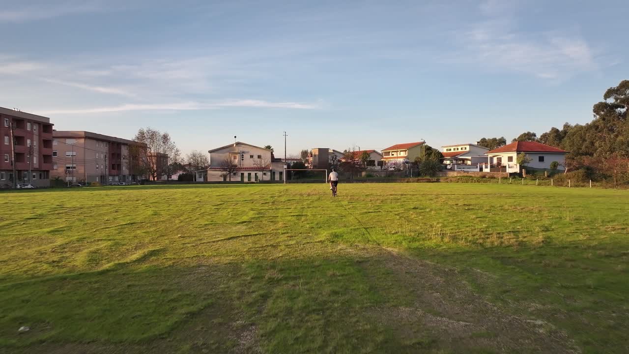 A drone follows a cyclist riding away across a green field with a white goalpost in view. Small four-floor suburban buildings frame the background under a bright sky.