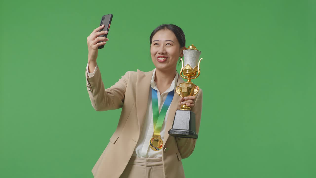스튜디오에서 녹색 화면 배경에서 스마트폰에서 비디오 통화에 금 트로피를 보여주는 금메달과 함께 슈트 아시아 비즈니스 여성