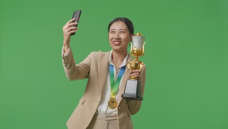스튜디오에서 녹색 화면 배경에서 스마트폰에서 비디오 통화에 금 트로피를 보여주는 금메달과 함께 슈트 아시아 비즈니스 여성