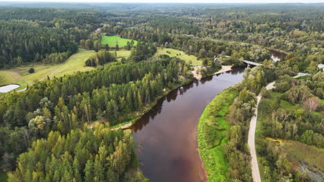 Scenic drone view of a winding river, lush forest, and green fields in Cesis, Latvia.