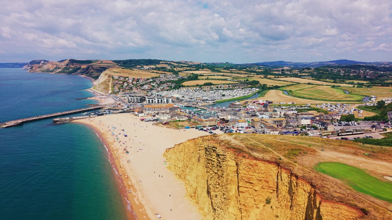 Ampie riprese aeree con drone che mostrano la spiaggia di ciottoli di West Bay, il porto, la città costiera e la dorata East Cliff sulla Jurassic Coast del Dorset, Inghilterra. Ampia inquadratura estiva di presentazione con mare azzurro