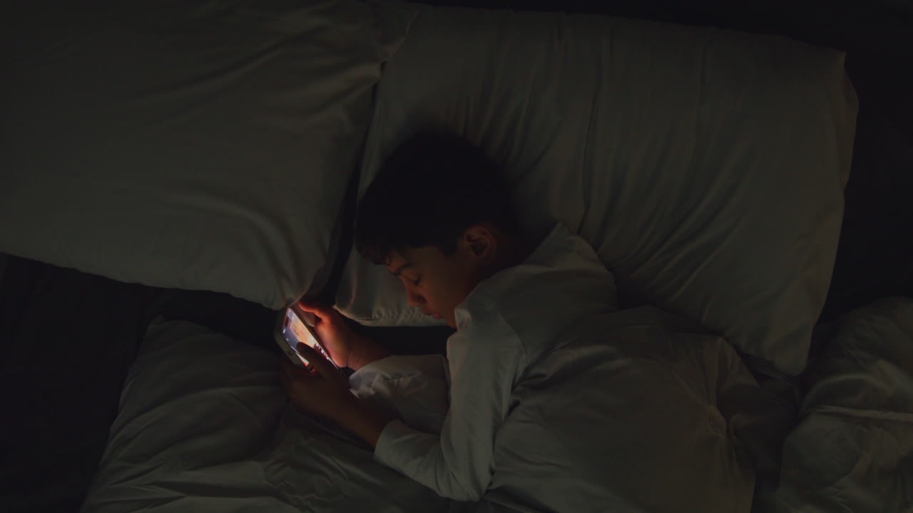 Young Boy In Bedroom At Home Lying In Bed Using Mobile Phone To Text Message At Night 2