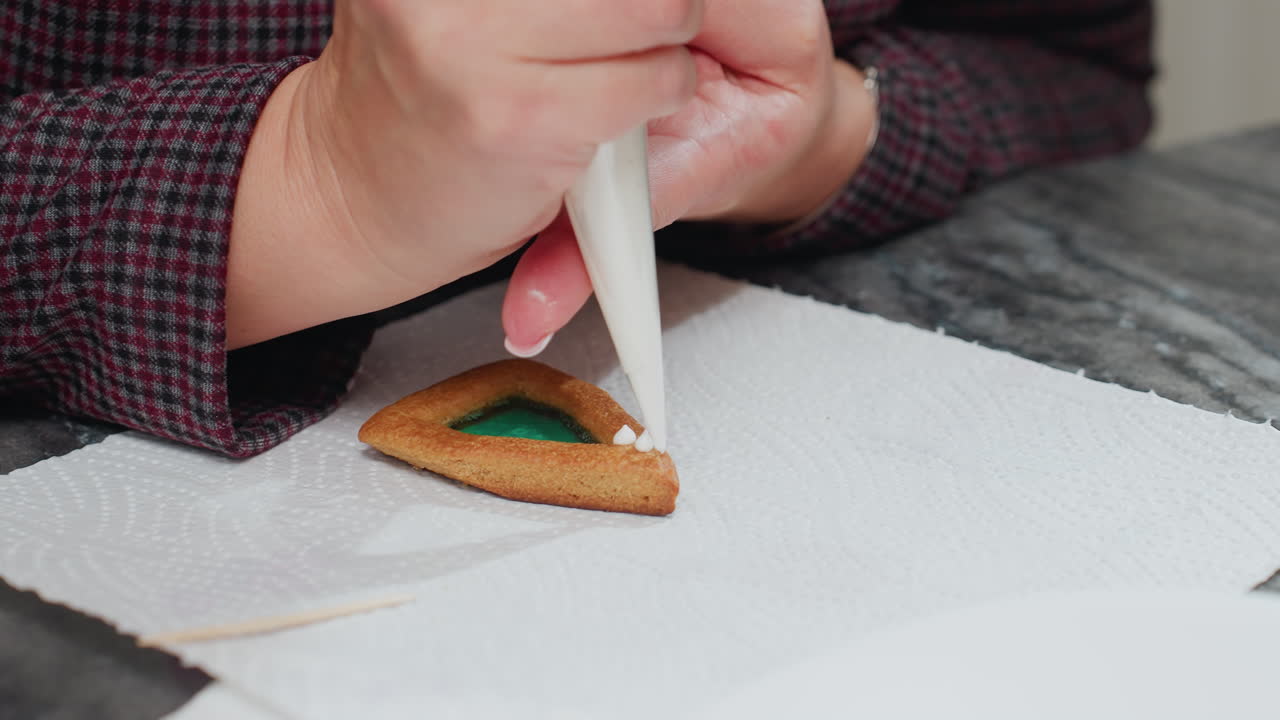 Close-up of baker using white piping bag to decorate cookie with green filling on tissue paper, toothpick nearby for precision as baker carefully applies icing to cookie edges