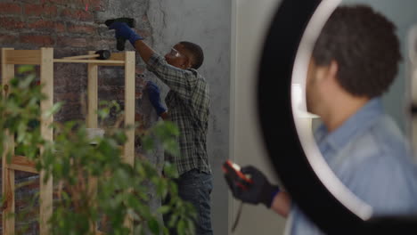 Black-man-points-laser-level-to-brick-wall-colleague-drills