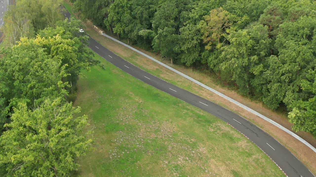Aerial view of white sports car driving on UTAC Mortefontaine race track surrounded by forest, France