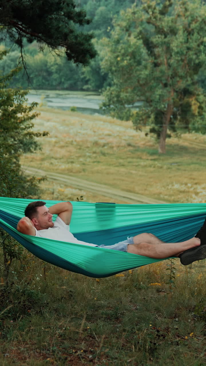Happy Caucasian man lies comfortably in a hammock hands behind his head. Relaxed person looks dreamily up swaying slowly in hammock. Nature at backdrop. Vertical video