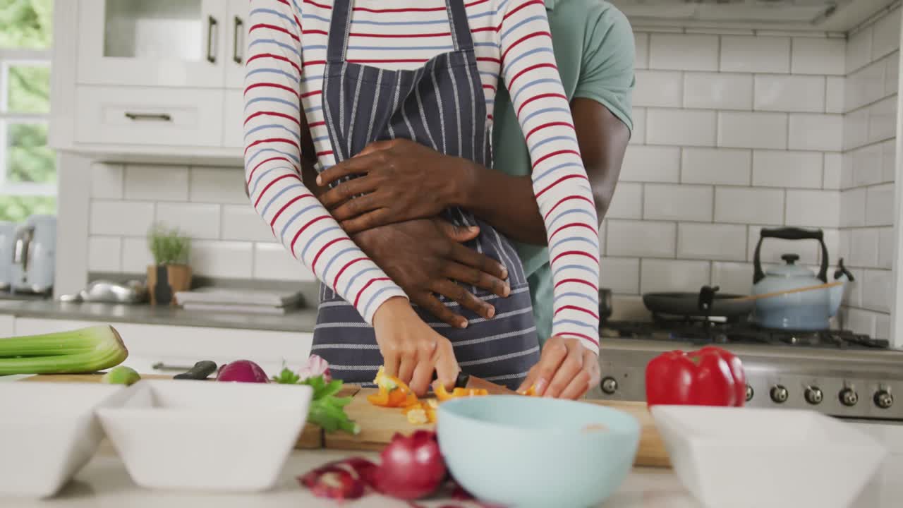 Diverse couple wearing blue apron embracing and cooking in kitchen