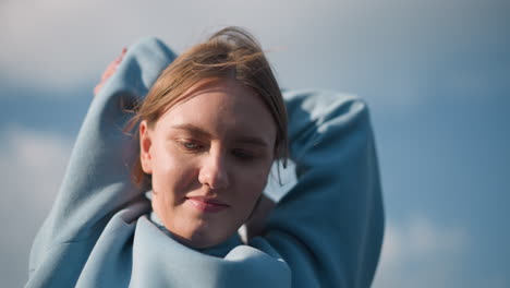 athlete in blue sweater engaging in fitness exercise, stretching arms overhead with a warm smile, sunlight highlights her face under a clear blue sky