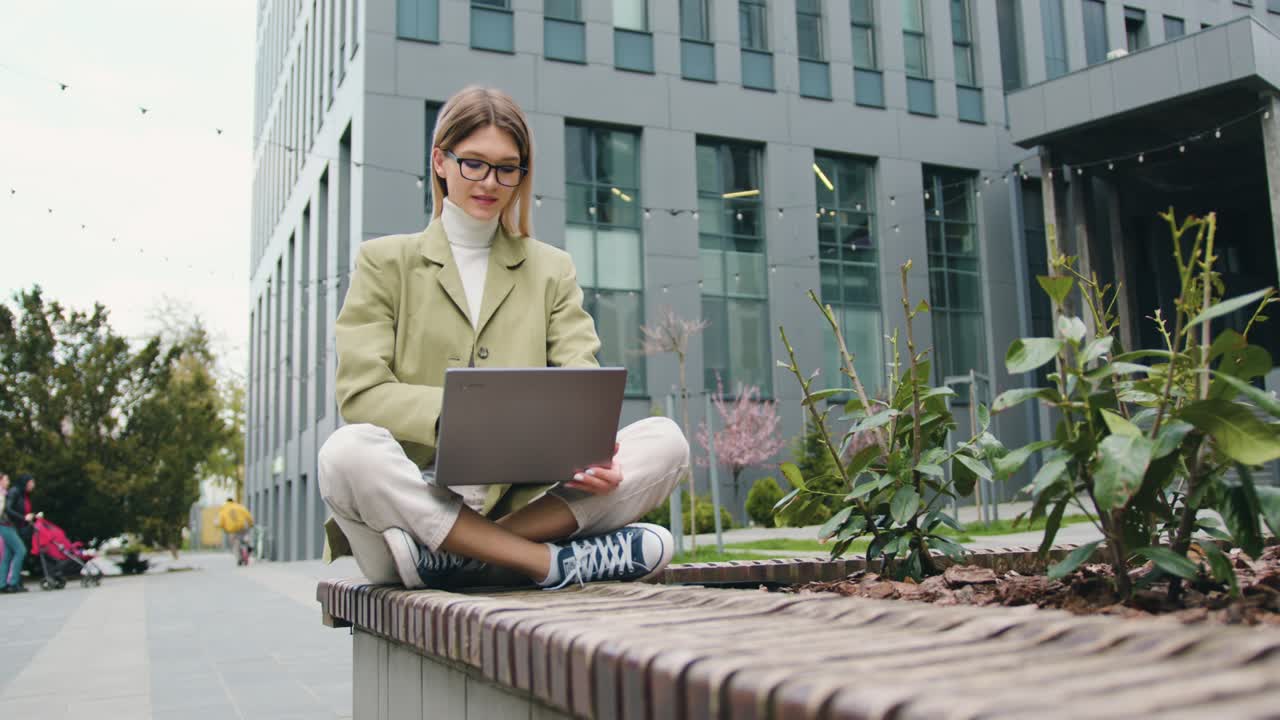 Confident young woman working outdoors sitting near office building typing on laptop using wireless computer for study