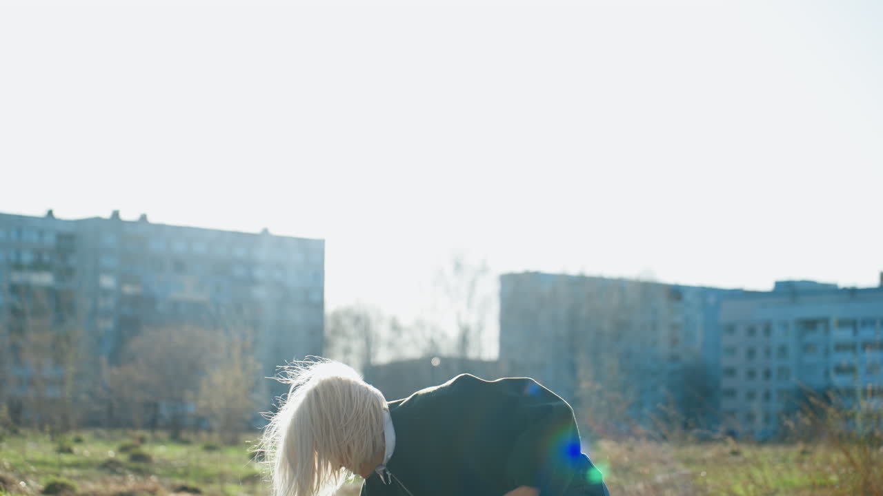 Blonde woman outdoors holding beige sweater in hands, wearing dark jacket and white shirt, sunlight shining from behind, urban residential buildings blurred in background, casual atmosphere