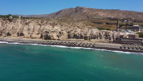 Aerial-of-greek-Island,-rocky-cliff-mountains-and-blue-water-waves-in-the-Mediterranean-Sea-on-white-beach-in-Santorini,-Greece