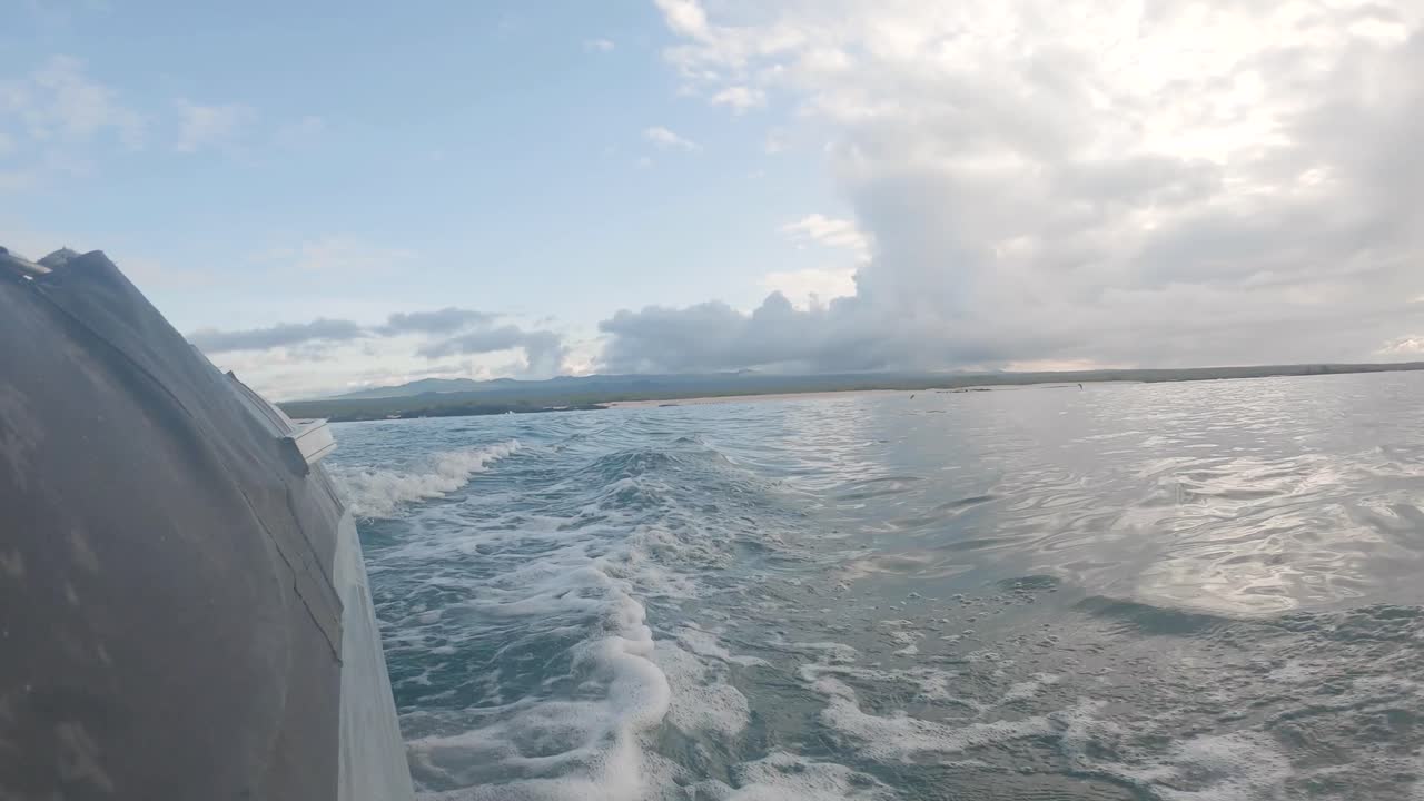 A bird flies close to the ocean while a boat moves away from an island. The panga creates waves on an ocean of the Galapagos islands