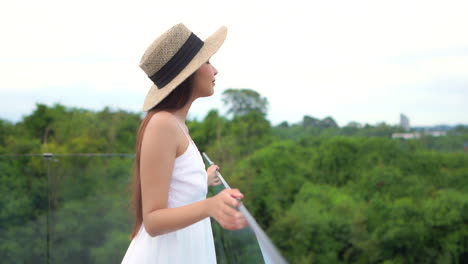 Asian-female-with-summer-hat-and-dress-on-luxury-modern-terrace-with-glass-fence,-thick-rainforest-in-background