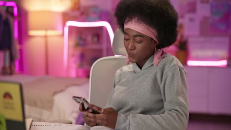 a young woman with curly afro hair, sitting at her desk, smiles and looks at her phone.