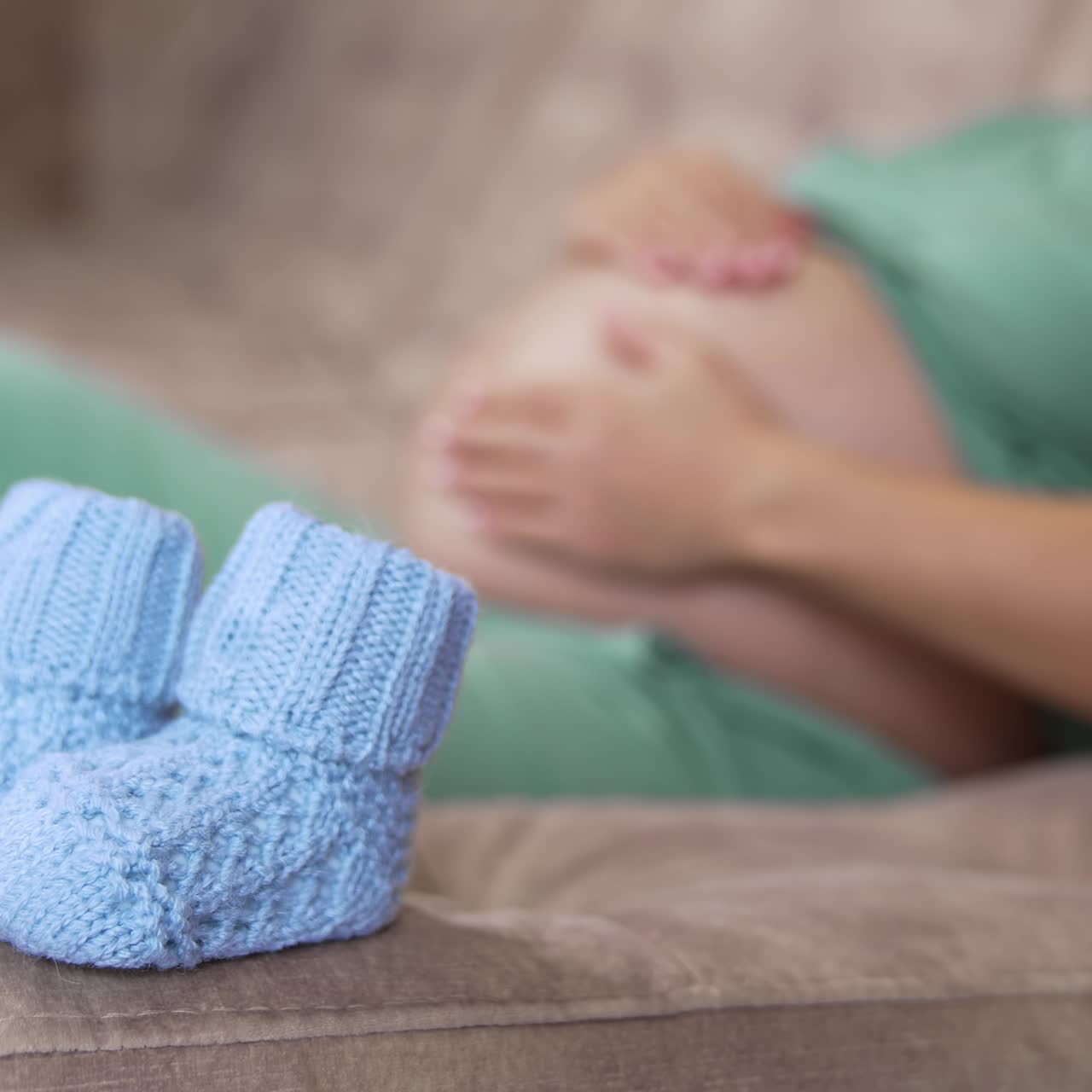 Little knitted socks for future baby. Blue baby socks on sofa on the blur background of a pregnant woman stroking her belly