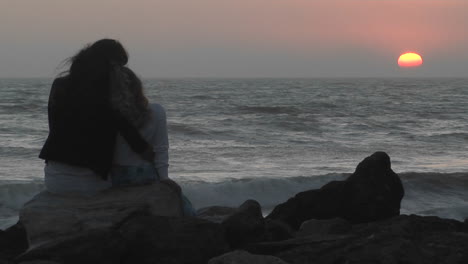 A-mother-and-her-young-daughter-sit-on-a-rock-facing-the-sea-at-golden-hour