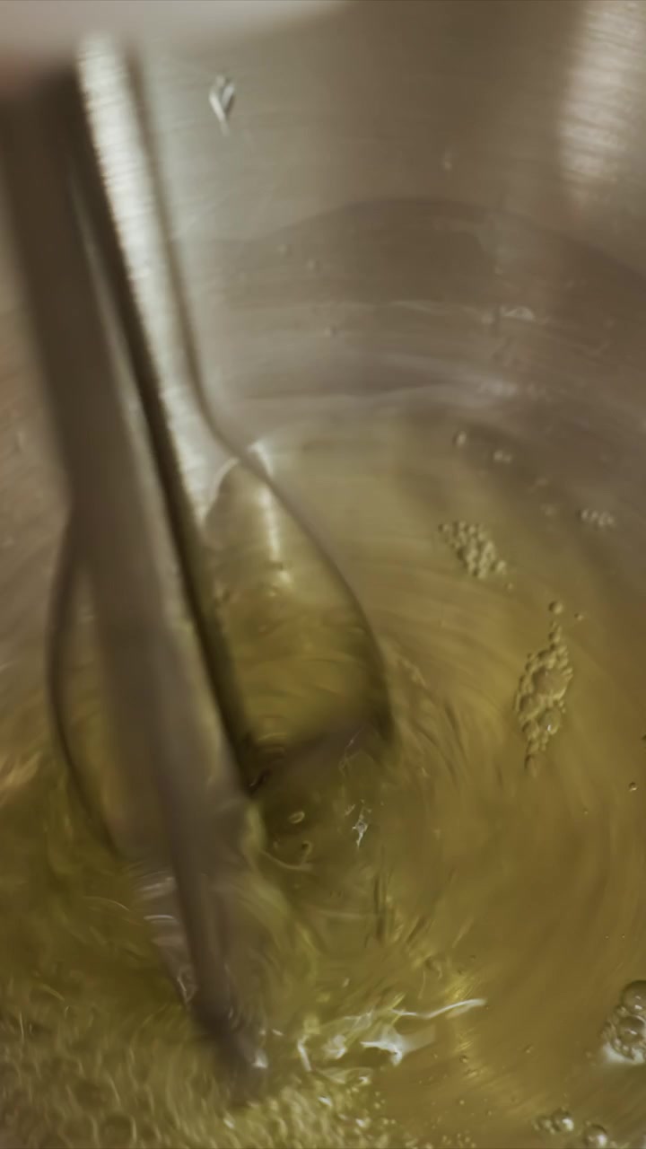 Primo piano di una frusta metallica che mescola un liquido giallo. Una mano oscura intermittentemente la vista, ma l'azione di mescolamento è evidente. Il liquido si agita e forma schiuma mentre la frusta si muove, catturato in un primo piano stabile.