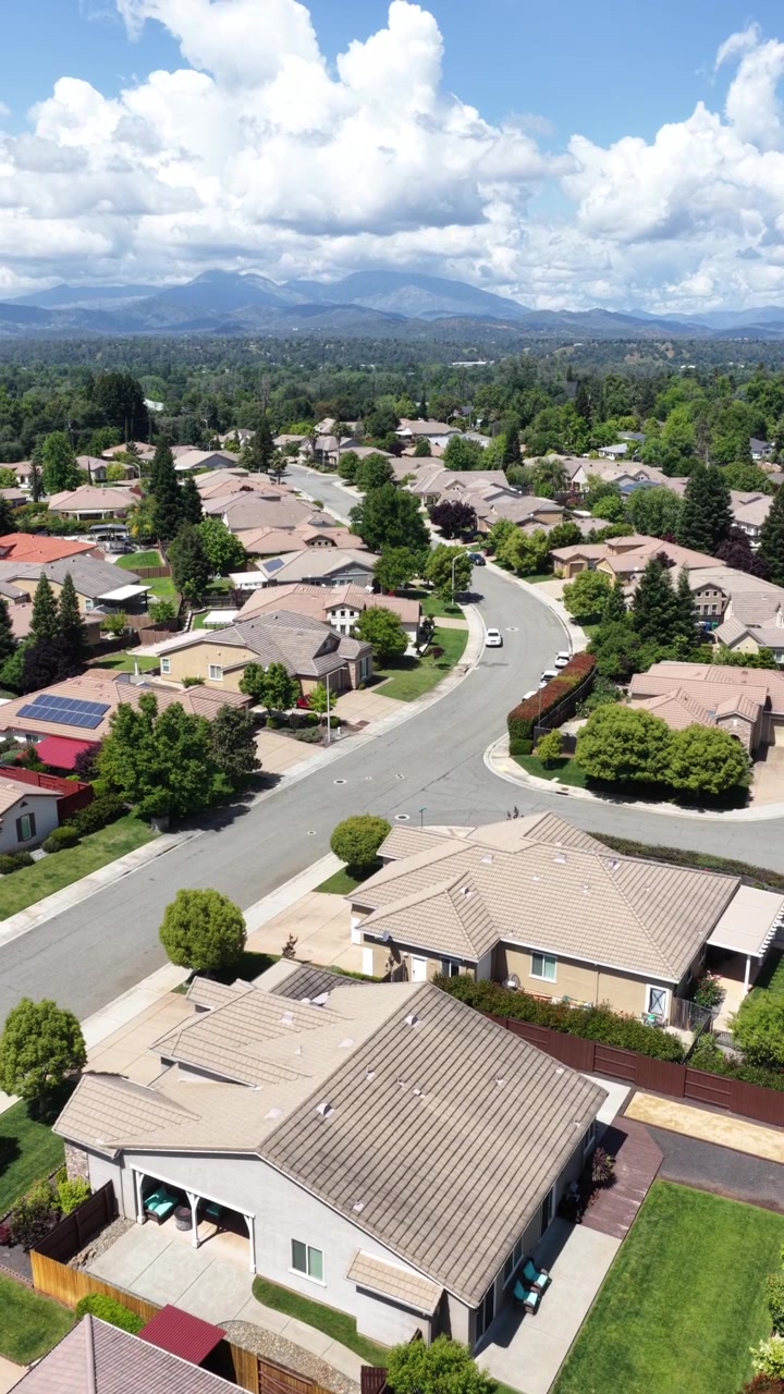 Techos de tejas rojas cubren casas suburbanas que bordean una calle sinuosa con paneles solares, además de pinos altos, estribaciones boscosas que se elevan hacia picos montañosos cerca de Redding, California. Toma vertical de dron aéreo.