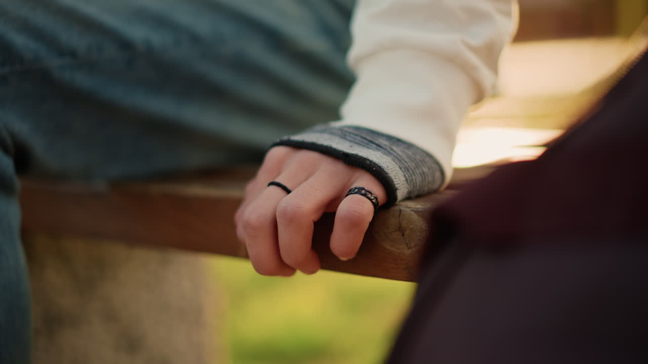 closeup hand near conversation foreground, finger curled on bench edge, blurred companion in foreground, dusk light and warm tones, indie musician waiting with nervous energy, cinematic tension