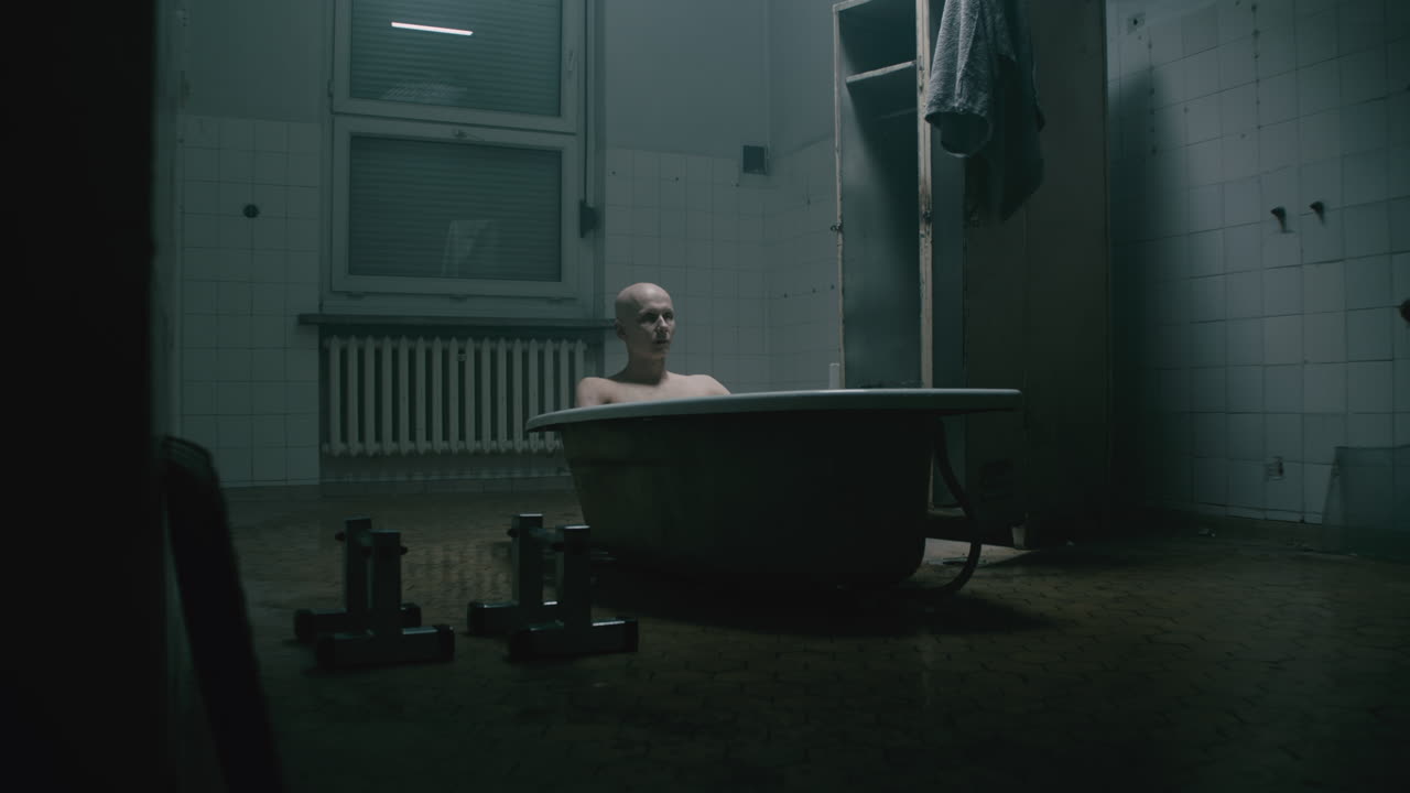Pale bald skinny man takes a bath in the creepy old bathroom at night