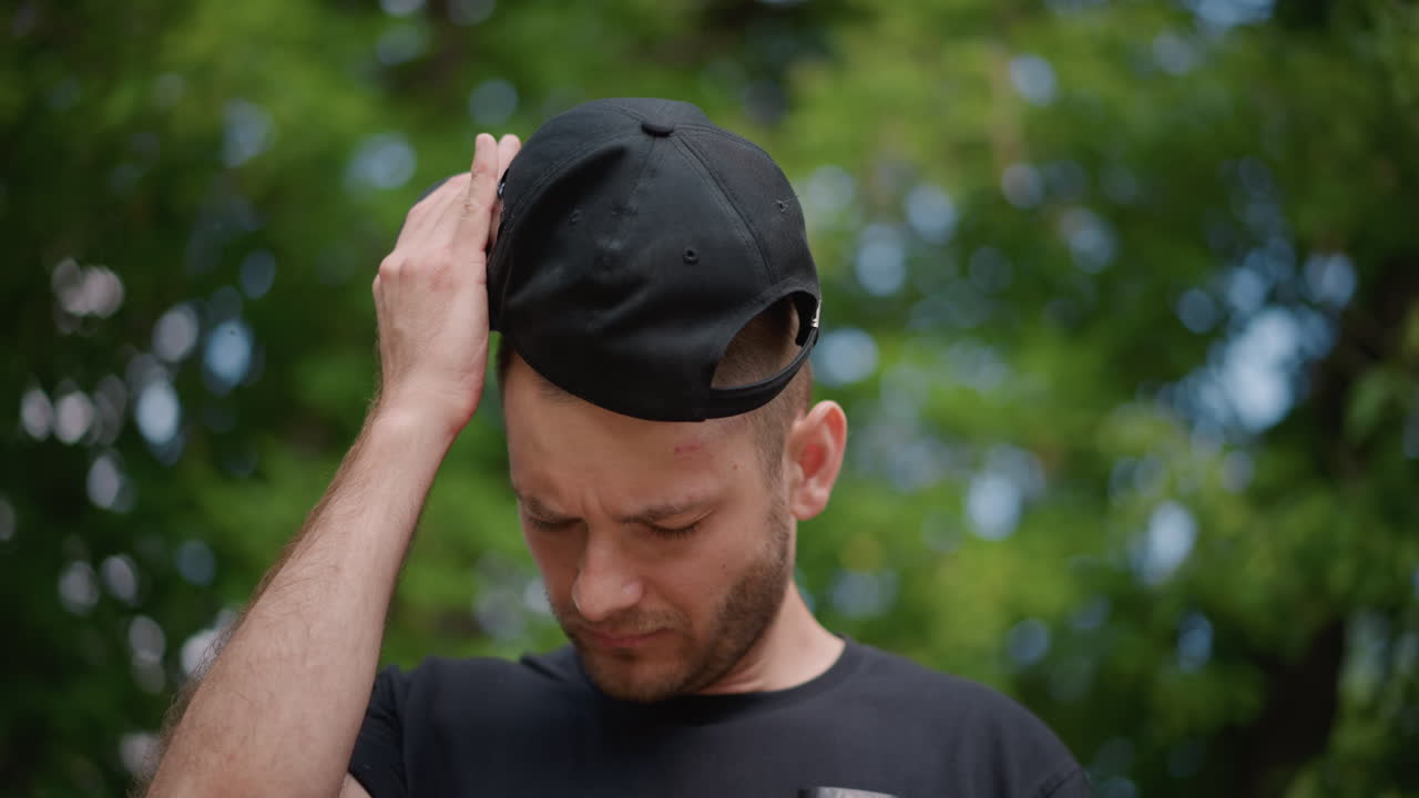Man In Park Taking Break, Man In Leafy Park Adjusts Cap Following Exertion During Leisure, Caucasian Male In Casual Attire Takes Moment To Recover After Exerting Himself Outdoors In Lush Park