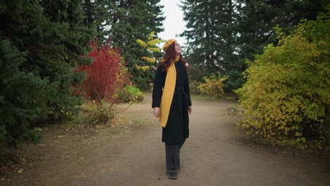 tourist with sunglasses and long red hair wearing yellow beret, matching scarf, and black coat, walking and smiling as she looks around vibrant garden with colorful autumn foliage